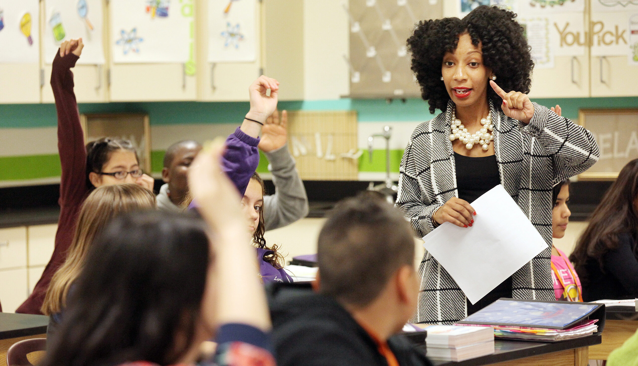 a teacher pointing to a student in a class with multiple hands raised in the air